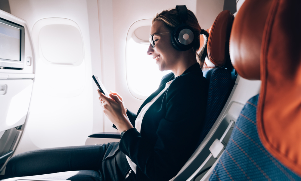 what to pack in your carry-on:Cheerful female passenger in headphones for noise cancellation watching online movie during intercontinental flight in cabin of aircraft