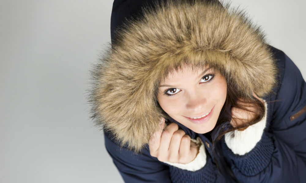 Sweet young girl in warm winter jacket with fur hood, looking up with a cute smile on her face, isolated on grey background.