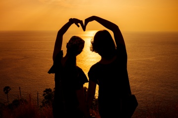 Silhouette photo of two girls arranged their arms in heart shape with sunset background