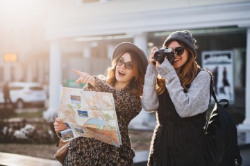 Happy travel together of two fashionable girls in sunny city centre. Young joyful women expressing positivity, using map, vacation with bags, camera, making photo, cheerful emotions, great mood