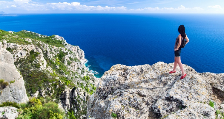 Travel girl stands on the edge of a high cliff above sea. Back view young female. Greece islands landscape. Travel Concept.