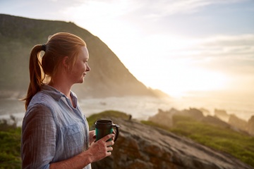 Young woman with light ginger hair and a fresh beauty, standing quietly with a flask of coffee in the early morning, looking out at a picturesque coastline