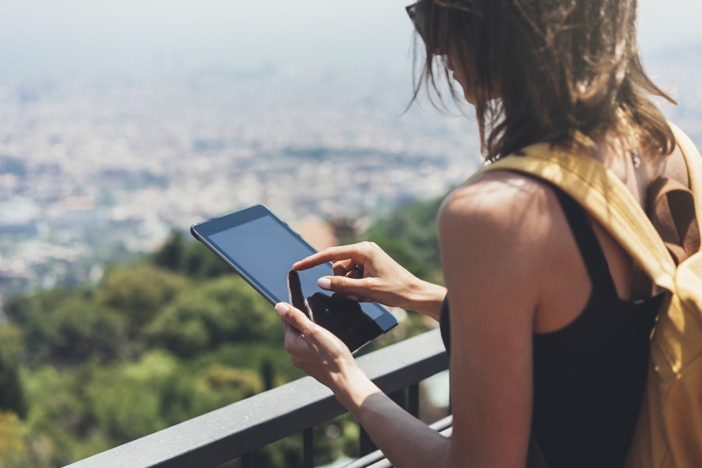 Girl with bright backpack planning travel plan. Traveler on background panoramic view of the city, binoculars. Mock up for text message. Barcelona Tibidabo. Female hands using tablet, holding gadget