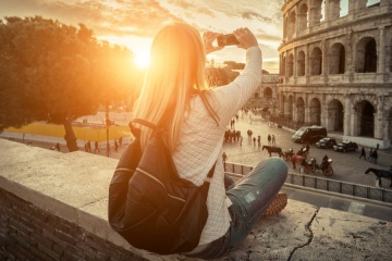 Woman tourist selfie with phone camera in hands near the Coliseum in Rome under sunlight and blue sky. Famous popular touristic place in the world.