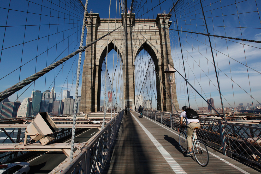 Bicyclist heading to Manhattan on the Brooklyn Bridge in morning light with blue sky and wispy clouds.