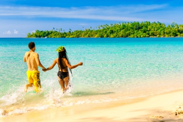 A loving couple, man and woman enjoying summer vacation on a tropical paradise beach with clear sea ocean water and scenic island on the horizon overgrown green palm on background blue summer sky