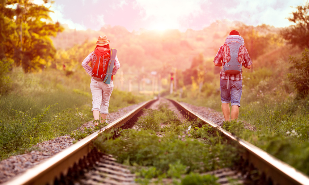 Two jolly Hikers Man and Girl in casual Travel Clothing with Backpacks walking along old Country Railroad with back light Sun and Forest on Background