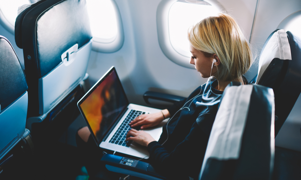  female passenger of airplane sitting in comfortable seat listening music in earphones while working at modern laptop computer with mock up area using wireless connection on board