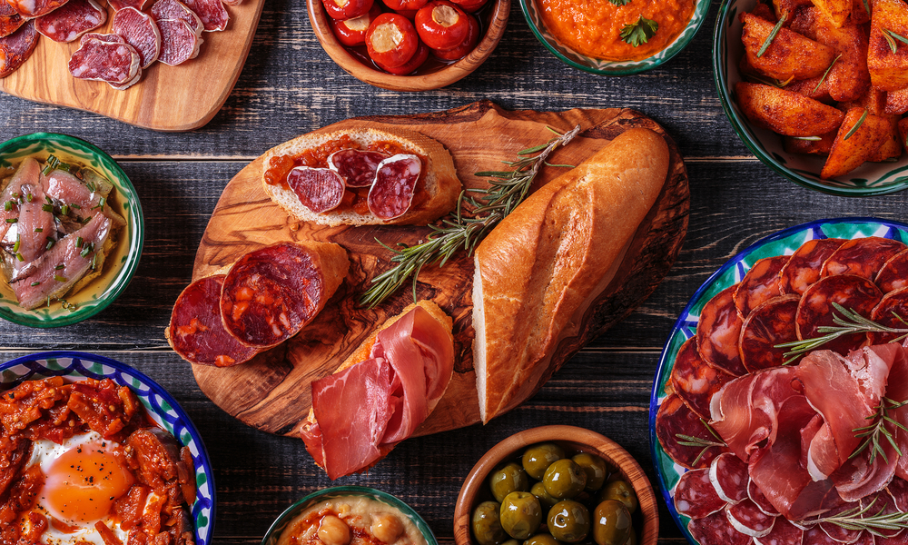 Typical spanish tapas concept. Concept include variety slices jamon, chorizo, salami, bowls with olives, peppers, anchovies, spicy potatoes, mashed chickpeas on a wooden table.