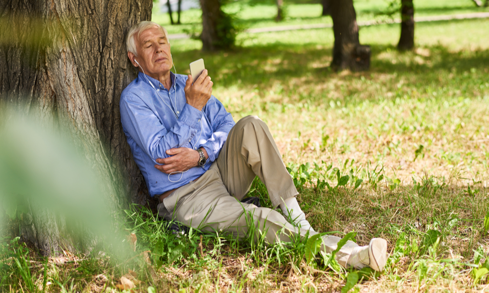 Portrait of modern senior man relaxing listening to music from smartphone in park under tree in shade