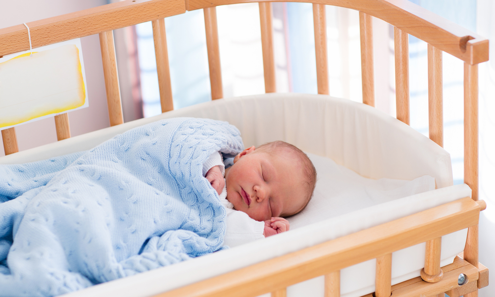 New born child in wooden co-sleeper crib. Infant sleeping in bedside bassinet. Safe co-sleeping in a bed side cot. Little boy taking a nap under knitted blanket.