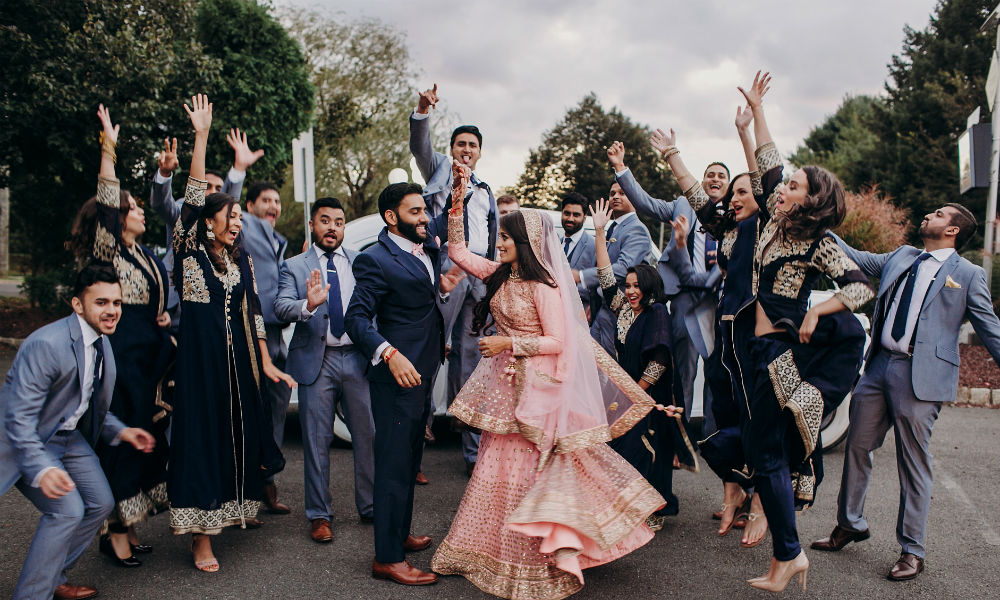 Indian groom dressed in traditional black suit and pretty bride in pink wedding dress with golden embroidery dance before a car together with their cheerful groomsman and bridesmaids