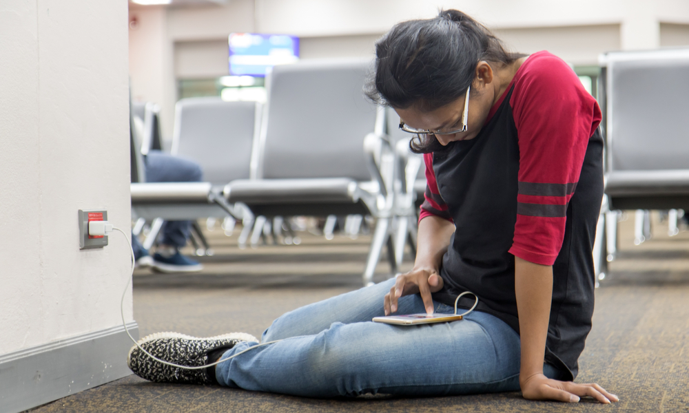 A young woman sit on floor and recharges the phone from the wall socket in the hall.