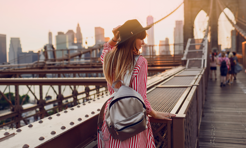  View from back of traveling woman in red dress, stylish black hat and silver bag pack enjoying amazing view from Brooklyn bridge in New York. 