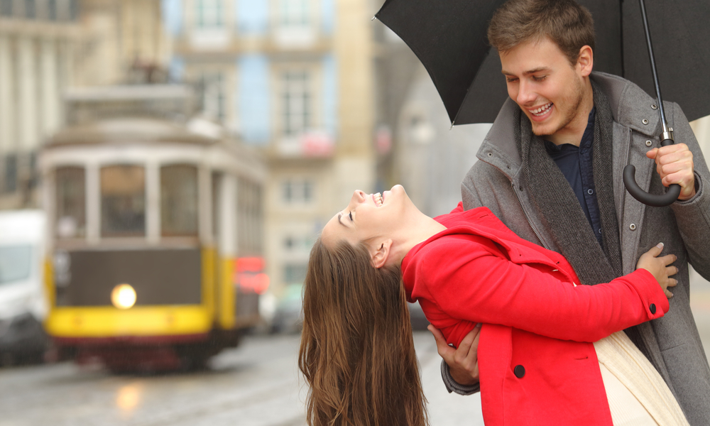 Happy playful couple in love dating joking and laughing in the street of an old town in a rainy day under an umbrella