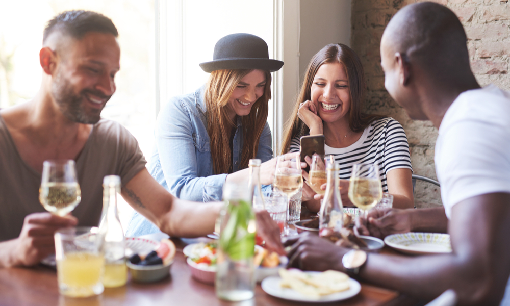 Group of four young friends having dinner in cozy cafe while women sharing new on phone