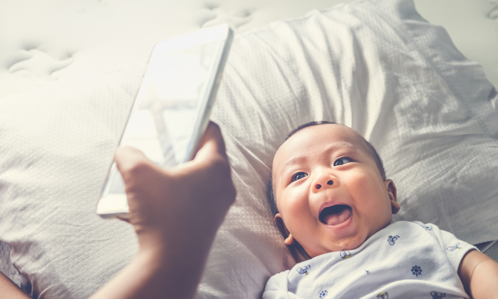 igh angle shot of happy mother taking picture her baby boy in bed with smart phone at home. 