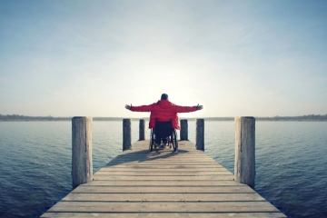 young man in wheelchair with wide opened arms enjoying his life at a lake in spring