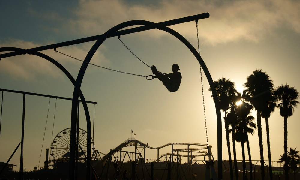 As the sun sets a silhouetted man works out on gymnast rings