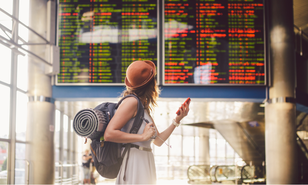 Beautiful young caucasian woman in dress and backpack standing inside train station or terminal looking at a schedule holding a red phone