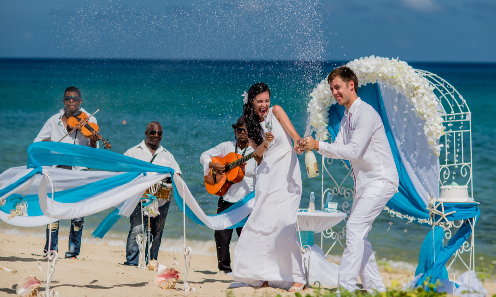 7 Winter Wedding Ideas That Will Help You Find the Perfect Spot to Say "I Do": Bride and groom are opening a champagne at a wedding ceremony with three musicians near caribbean coastline