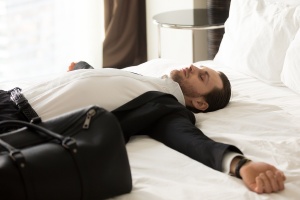 How to overcome jet lag: Exhausted young businessman laying with outstretched arms on bed next to small suitcase in hotel room