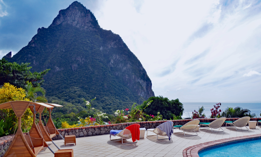 The Best Winter Getaways Where You Can Forget the Cold: Swimming pool, lounges with bright beach towels, and shaded swings on stone deck lined with tropical flowers overlooking St. Lucia island's Petit Piton and the Caribbean sea against a cloudy blue sky.