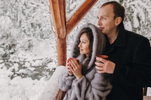 Stylish couple holding hot tea in cups and looking at winter snowy mountains from wooden porch