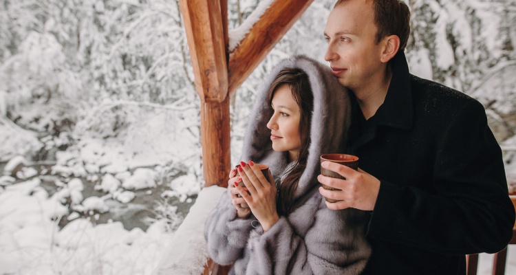 Stylish couple holding hot tea in cups and looking at winter snowy mountains from wooden porch