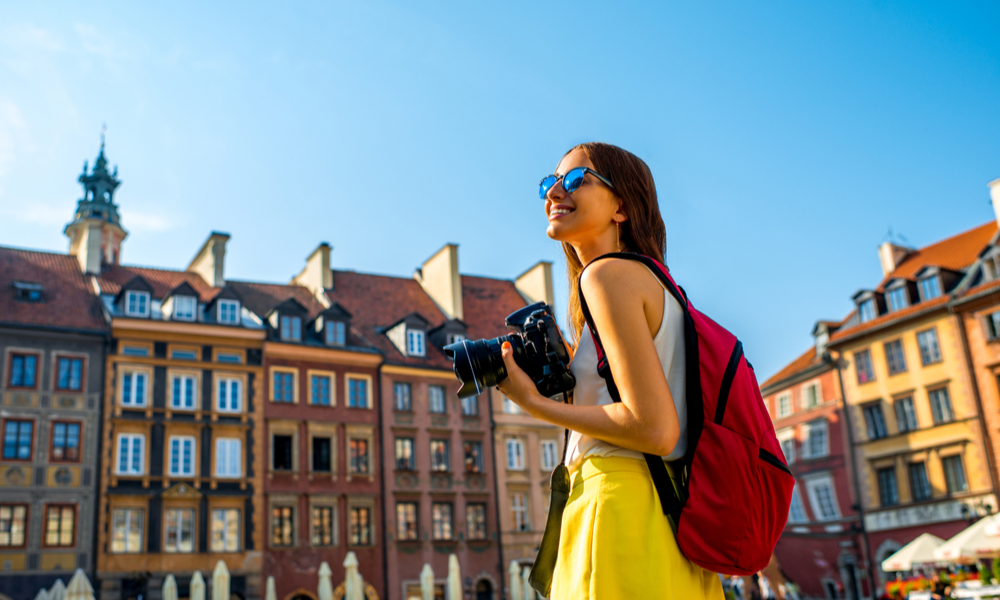 How To Plan a Vacation: Female young traveler with backpack and photocamera in the old town market square in Warsaw, Poland