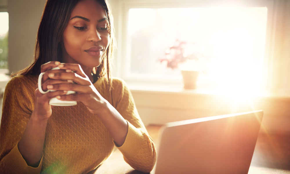 Tips for alternatives to parking at the airport: Adult woman smiling sitting near bright window while looking at open laptop computer on table and holding white mug