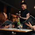 Cheerful two young women sitting at cafe holding menu card giving order to waiter. Young woman placing order to a waiter at restaurant while sitting with her friend.