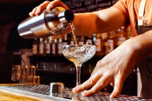 Male bartender is making cocktail pouring alcohol from shaker to glass at bar background.